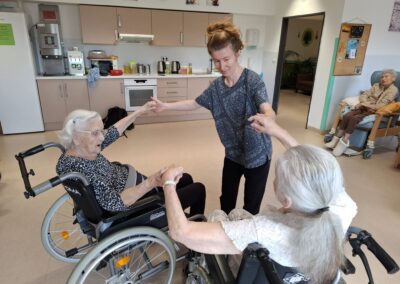 Lektorka tančí vsedě s klientkou na vozíku, druhá seniorka se k nim přidává
