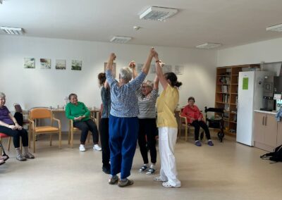 Skupina seniorů tančí ve stoje s rukama nad hlavou, ostatní sedí kolem