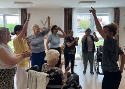 Senioři a seniorky zvedají ruce při taneční hodině v domově, lektorka tančí s nimi