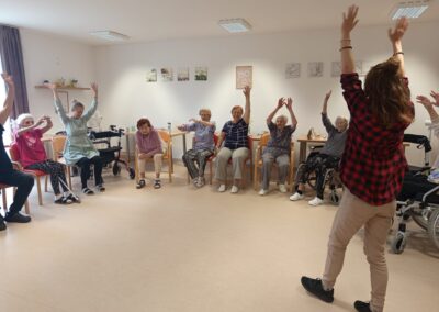 Lektorka zvedá ruce uprostřed kruhu sedících seniorů