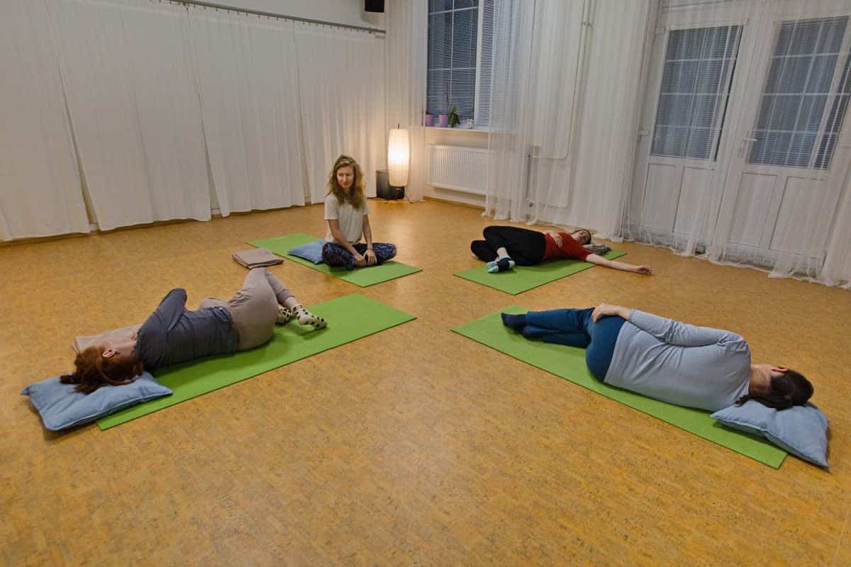 Dvě ženy odpočívají na bocích na podložkách, lektorka sedí v tureckém sedu